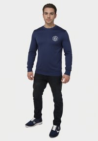 Camiseta de manga larga azul marino con cuello redondo y detalle de logo blanco en el lado izquierdo del pecho, combinada con jeans oscuros y zapatillas azul marino.