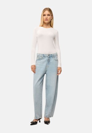Giovane donna con maglia bianca a maniche lunghe, jeans larghi azzurri e scarpe nere con tacco, in piedi davanti a uno sfondo semplice.