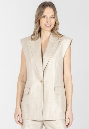 Kobieta w beżowej bezrękawnikowej marynarce i dopasowanych spodniach, uśmiechająca się, z luźnymi falującymi włosami na tle jednolitego jasnego tła.