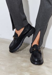 Black loafers made of leather with a textured finish, featuring a gold-tone buckle and a chunky, rugged sole. Worn with gray pants.
