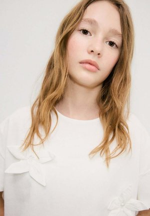 Giovane donna con capelli castano chiaro che indossa una camicia bianca con dettagli di fiori in tessuto su uno sfondo semplice.