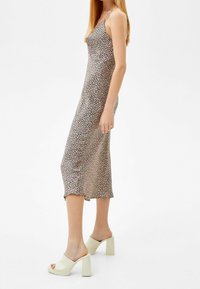 Kobieta w średniej długości kwiecistej sukience na ramiączkach z cienkimi paskami i beżowych szpilkach z odkrytymi palcami, stojąca bokiem na jednolitym tle.