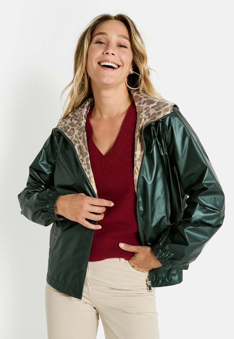 Mujer sonriente con chaqueta verde oscuro con forro de estampado de leopardo sobre un suéter rojo y pantalones beiges, posando con una mano en el bolsillo.