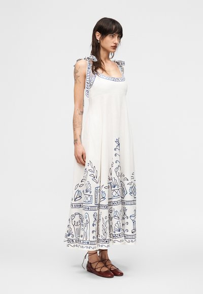 Γυναίκα με τατουάζ φοράει λευκό αμάνικο φόρεμα με μπλε διακοσμητικά σχέδια και τιράντες με φιόγκους, συνδυασμένο με καφέ δερμάτινα παπούτσια με κορδόνια, στέκεται πλαγίως.