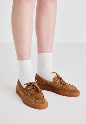 Bruine suede schoenen met een rubberen zool, voorzien van veters, metalen oogjes en gedragen met witte crew sokken.