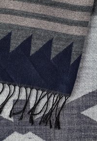 Tissu tissé avec frange noire, présentant des motifs triangulaires bleu foncé et des rayures horizontales en tons gris et beige.