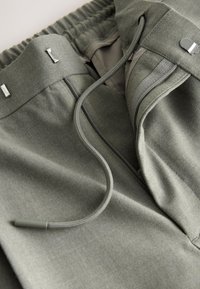 Gros plan sur un pantalon vert avec taille élastique, cordon, braguette zippée et fermetures à crochets et barres métalliques.