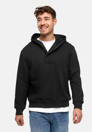 Homme souriant portant un sweat à capuche noir sur une chemise blanche et un jean bleu, debout devant un fond uni.