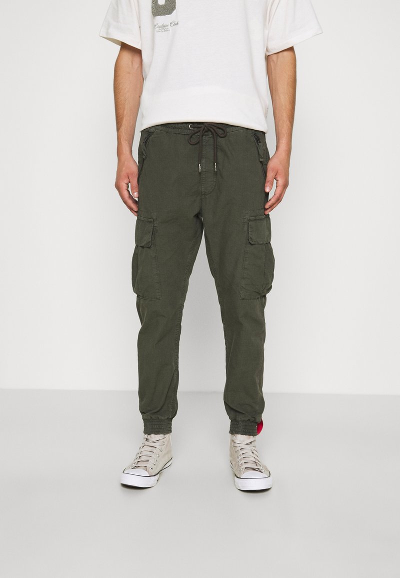 Olijfgroene cargobroek van stof met een trekkoord in de taille, taps toelopende benen en meerdere zijzakken. Gecombineerd met beige hoge sneakers.