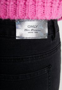 Primer plano del bolsillo trasero de unos jeans de mezclilla negros con la etiqueta metálica ONLY Blue Denim y un suéter de punto rosa encima.