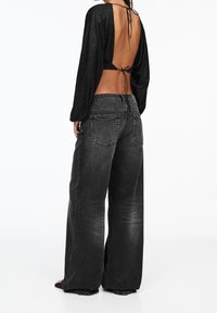Schwarzes Langarmoberteil mit offener Rückseite und Bindedetail, kombiniert mit lockeren, dunkelgrauen, weiten Jeans. Sanfte Texturen, minimale Details.