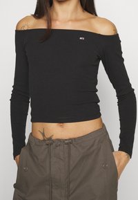 Schwarzes, geripptes, schulterfreies Langarmoberteil mit tailliertem Design. Kleines Logoakzent auf der Brust. Kombiniert mit leichten, olivgrünen Hosen.