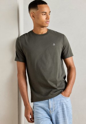 Homme portant un t-shirt vert foncé avec un petit logo blanc sur la poitrine et un jean bleu clair, regardant sur le côté avec une main dans sa poche.