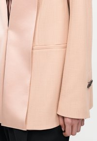 Κοντινό πλάνο ενός μπεζ σακακιού ραμμένου στα μέτρα με υφασμένη τσέπη και μανίκι που διαθέτει διακοσμητικό μεταλλικό κουμπί.