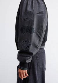 Zwarte leren bomberjack met geborduurde patches op de mouwen, geribbelde manchetten en een aansluitend ontwerp. Matte textuur.