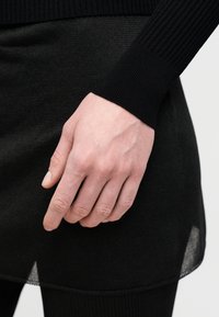 Μαύρο πλεκτό μανικετόπλεχτο πουλόβερ με ριμπ μανσέτες και ένα διαφανές κάτω στρώμα μιας μαύρης φούστας, που αναδεικνύει την υφή και τις λεπτομέρειες σχεδίασης.