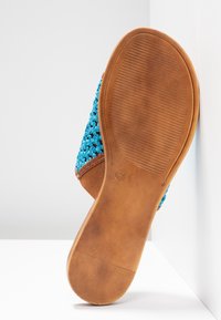 Semelle d'une sandale plate marron avec une semelle en caoutchouc nervurée et une tige tissée bleue, présentée verticalement contre un fond blanc en angle.