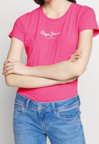 Pink cotton t-shirt with short sleeves and a round neckline, featuring a white logo text "Pepe Jeans" on the chest, paired with blue jeans.