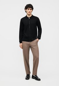 Mann stående, iført svart hettegenser med glidelås, beige bukser og svarte pensko mot en ensfarget hvit bakgrunn.