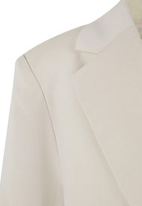 Détail d'épaule et de revers d'un blazer beige clair, tissu lisse avec couture ajustée visible sur un côté.