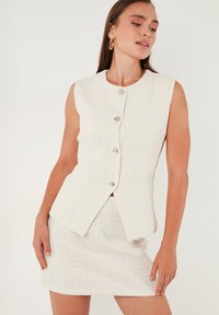 White tweed sleeveless blazer with a fitted cut and round neckline, featuring matching mini skirt. Silver button accents with a textured finish.
