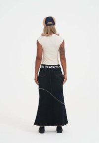 Falda maxi de mezclilla con dobladillo deshilachado y detalle curvado, combinada con una camiseta blanca ajustada. Cabello en ondas sueltas, usando una gorra bicolor.
