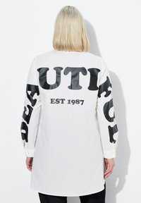 Maglietta bianca a maniche lunghe con testo oversized nero che recita "BEAUTIFUL" sulle maniche e "EST 1987" sul retro. Tessuto morbido, orlo curvo.