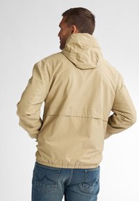 Jachetă bej cu glugă, cu o croială lejeră, tiv elastic și țesătură texturată. Are un umeră posterior și detalii de design simple. Este purtată cu blugi albaștri.
