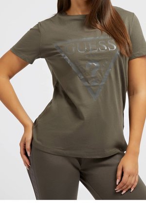 Mujer con camiseta verde oliva de Guess con logo de triángulo y pantalones a juego, mano apoyada en la cadera, sobre un fondo liso.