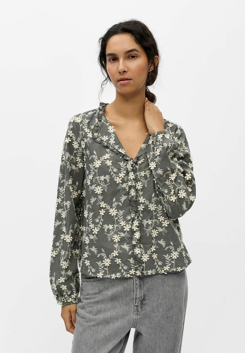 Femme portant un chemisier gris brodé de fleurs avec col à volants et manches longues, associé à un jean taille haute gris clair.
