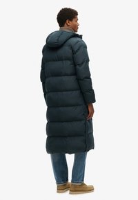 Lange donkergroene pufferjas met een capuchon, met brede horizontale secties, een hoge kraag en zijzakken. Gedragen over een blauwe spijkerbroek en beige schoenen.