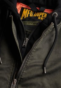 Zielona, pikowana kurtka z czarnym kapturem i podszewką. Posiada srebrny zamek błyskawiczny oraz metkę z napisem "MFG. SUPERDRY" w kolorze żółtym. Materiał o teksturowanej fakturze.