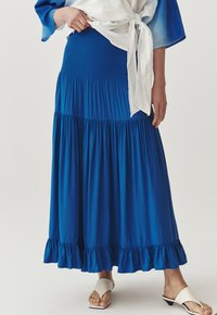 Gonna maxi blu a balze con vita arricciata, orlo con volant, realizzata in tessuto morbido. Indossata con un top leggero annodato e sandali bianchi con punta aperta.