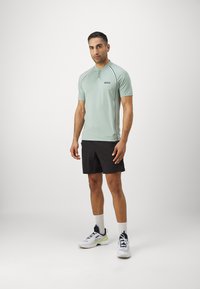 Miętowozielona koszulka polo odprowadzająca wilgoć z czarnymi akcentami, krótki rękaw i kołnierzyk; zestawiona z czarnymi szortami sportowymi i białymi trampkami.