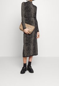 Robe noire à manches longues avec un léger motif texturé, longueur mi-mollet. Un sac à main en beige tissé et des mocassins noirs épais complètent le look.