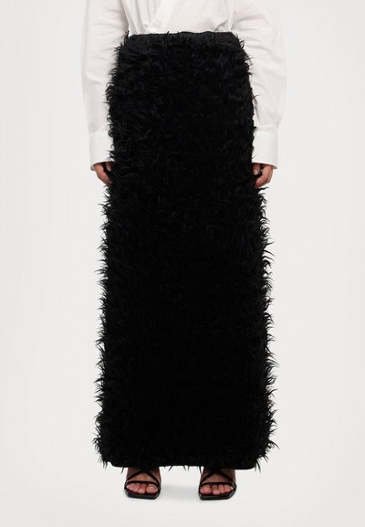 Jupe maxi noire en fourrure avec une surface texturée et shaggy. Portée avec une chemise blanche à boutons et des sandales noires à lanières.