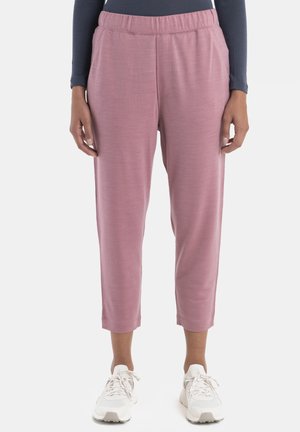 Personne portant un pantalon rose raccourci avec une ceinture élastique et des baskets blanches, debout devant un fond uni.