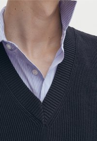 Mörk ribbad stickad tröja med V-krage, lagd över en ljusblå och vit randig skjorta med knappar. Kragen är något uppvikt.