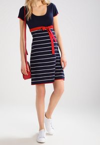 Kvinna klädd i en marinblå knälång klänning med vita ränder och röd trim, röd midjeband, bärande på en röd axelväska och vita sneakers.