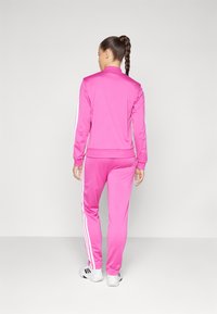 Pink athletic tracksuit featuring white side stripes, fitted design, and high collar. Made of smooth, stretchable fabric.