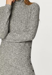 Graues Strickkleid mit hohem Kragen, gerippter Textur und seitlichen Knopfdetails. Die taillierte Silhouette betont die Körperform.