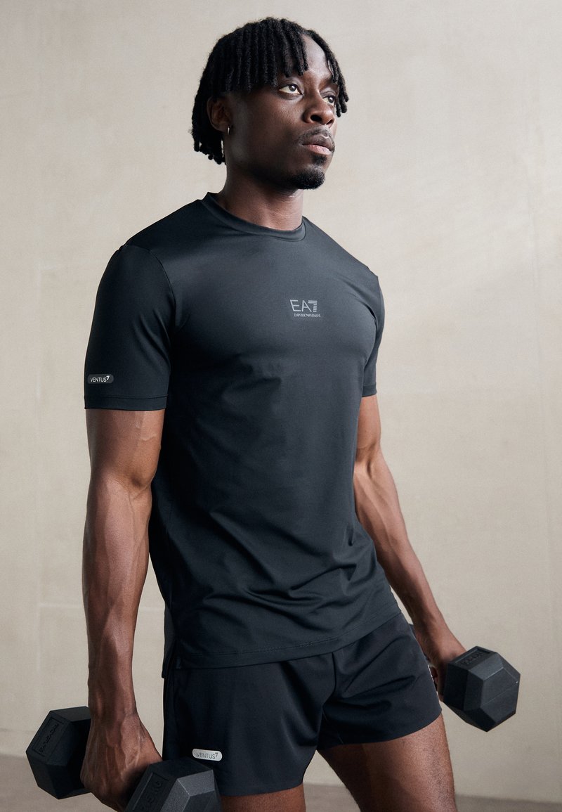 T-shirt de sport noir à manches courtes avec un logo, assorti à un short noir. Le mannequin tient deux haltères hexagonaux, mettant en valeur des bras musclés et une silhouette athlétique.