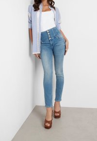 Jeans de mezclilla azules de cintura alta con cinco botones en la parte frontal y corte ajustado, combinados con una camiseta de tirantes blanca y una camisa a rayas azul, y tacones de plataforma marrones.