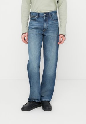 Lichtblauwe denim jeans met een rechte pijp, voorzien van vijf zakken en een leren patch op de achterkant van de tailleband, gecombineerd met zwarte schoenen.