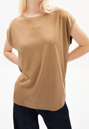 Blond persoon die een kort mouwzaam, loszittend t-shirt in camelbruin draagt en donkerblauwe broek, staand met de handen achter de rug.