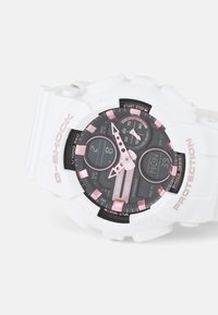 Montre G-Shock blanche avec un cadran noir et des accents roses, affichages numériques et analogiques, et bracelet en caoutchouc texturé pour la durabilité.