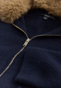 Giacca con zip blu navy con colletto in pelliccia sintetica, caratterizzata da una cerniera dorata e tessuto texturizzato. L'etichetta mostra il marchio "NEXT" e la taglia "S".