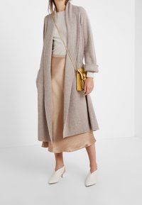 Beżowy kardigan z prążkowanym wzorem, jasnoszara bluzka, satynowa beżowa spódnica i białe modne klapki. Złota torebka na ramię z łańcuchowym paskiem, gładka faktura.