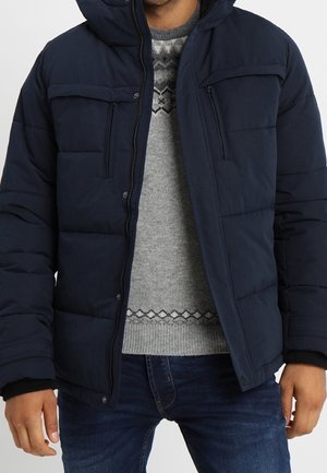 Mann trägt eine marineblaue gesteppte Winterjacke über einem grauen, gemusterten Pullover und einer blauen Jeans, steht mit entspannten Armen.