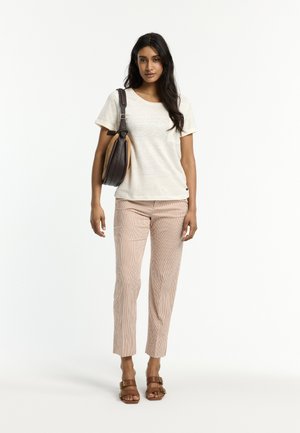 Frau trägt ein weißes Kurzarm-Shirt, hellrosa gestreifte Hosen, braune Sandalen und trägt eine dunkelbraune Umhängetasche, steht vor weißem Hintergrund.